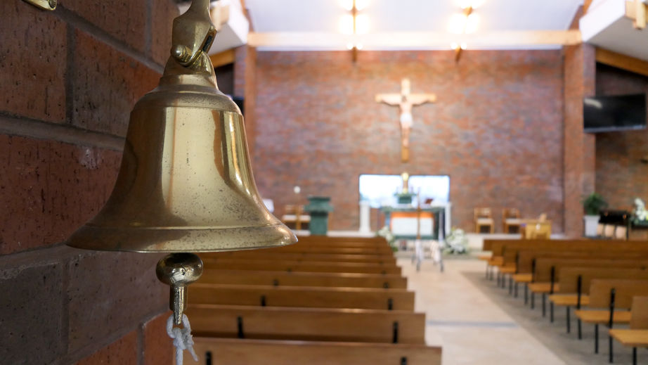 A brass bell hangs inside a church with wooden pews, facing the altar and a crucifix on a brick wall.