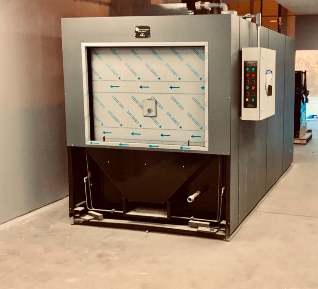Industrial heating chamber with control panel, featuring metal walls and a vented section below, situated on a concrete floor.