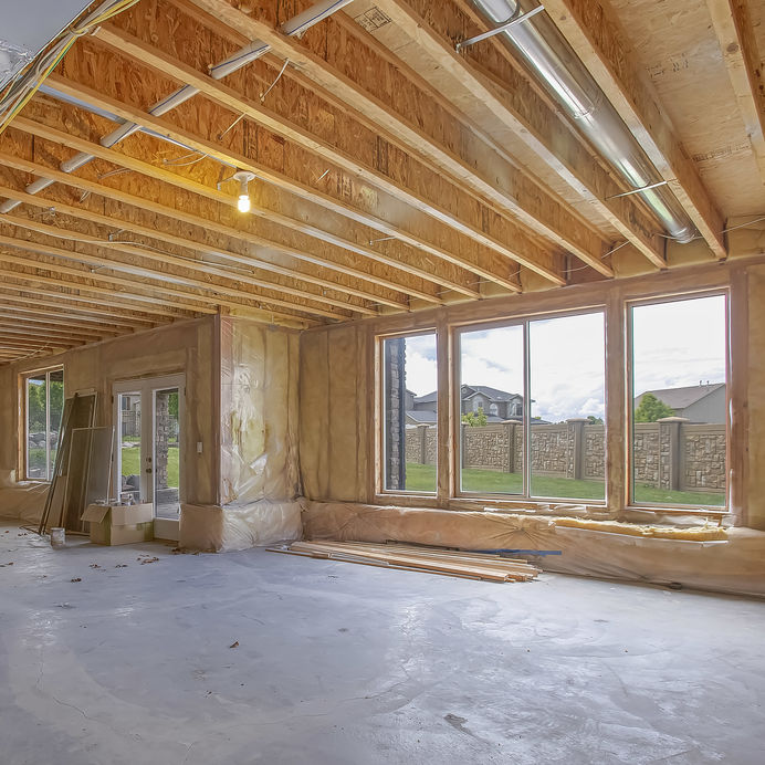 Unfinished room with exposed wooden ceiling beams, large windows, and a cement floor, overlooking a grassy outdoor area.