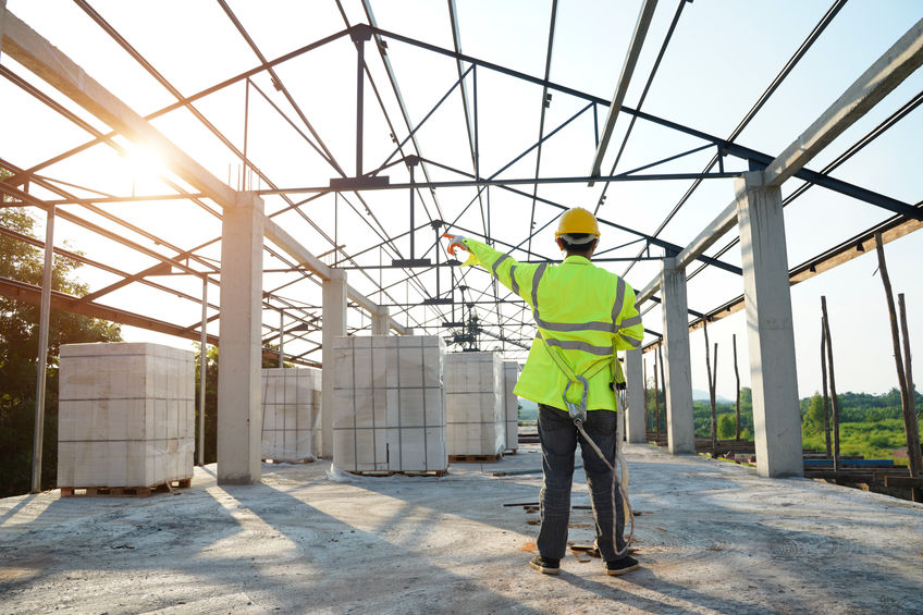 A construction worker in a high-visibility jacket and hard hat points at structural steel beams on a building site under sunlight.