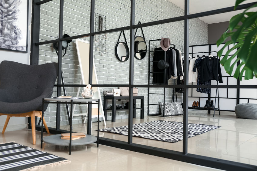 Modern room with glass partition, gray chair, round table, rugs, and clothing rack. Light brick wall and potted plant.